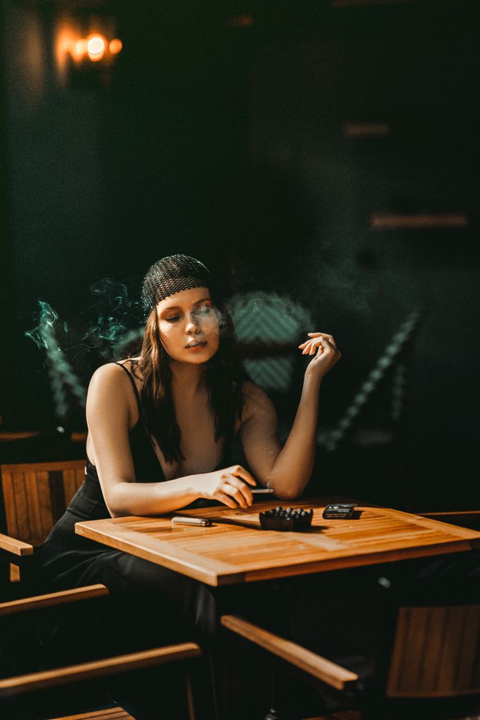 Stylish young woman smoking and relaxing in a dimly lit lounge environment, highlighting a mood of relaxation and elegance.