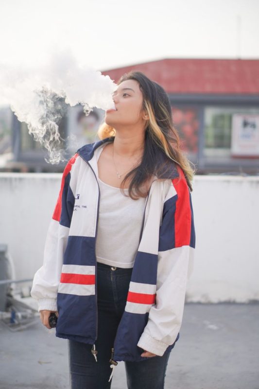 A young woman in a stylish jacket vaping outdoors under daylight.